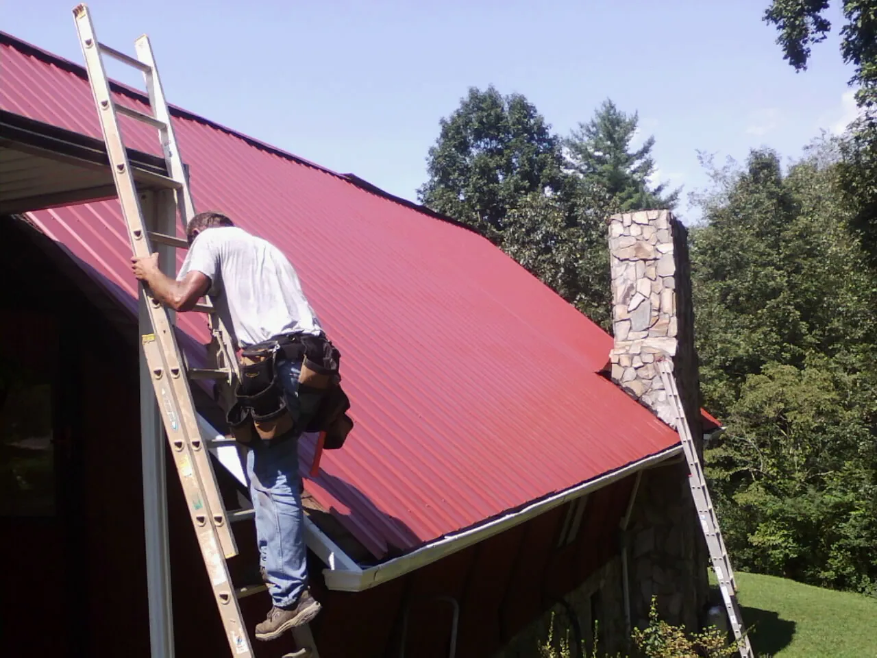 Roofer on a ladder beside a red metal roof and stone chimney — Soffit & Fascia Repair by Roofing Brothers in Smithmill